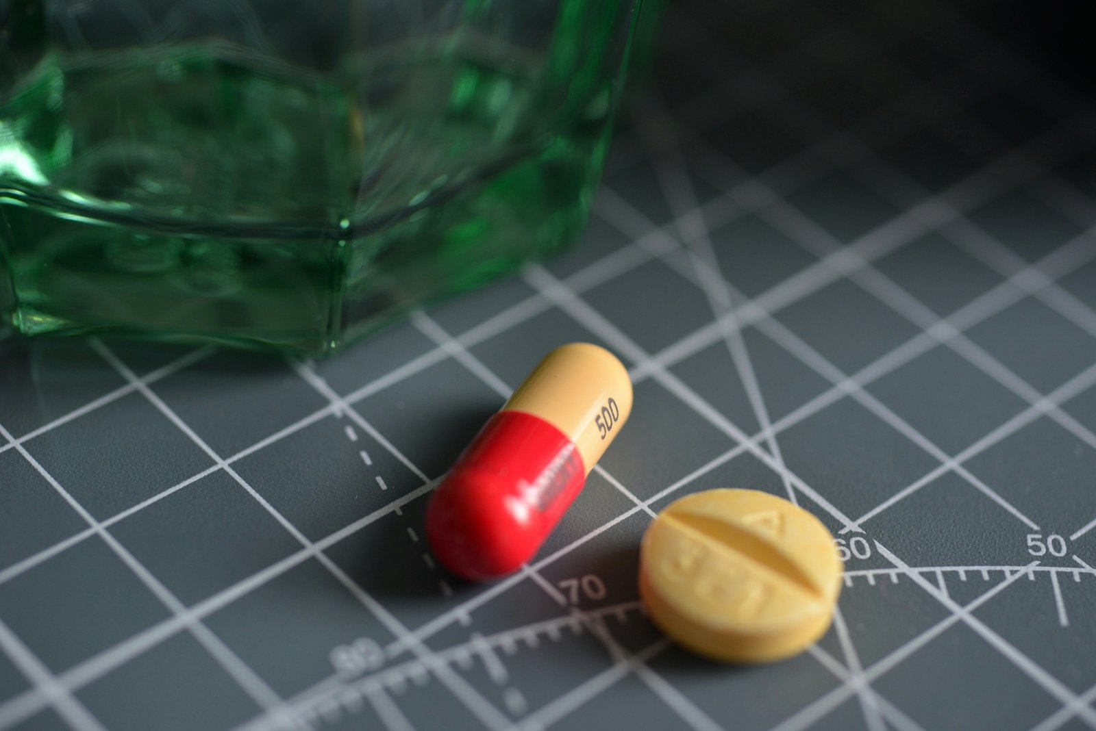 A red and yellow capsule and a yellow tablet on a dark grid surface beside green glass
