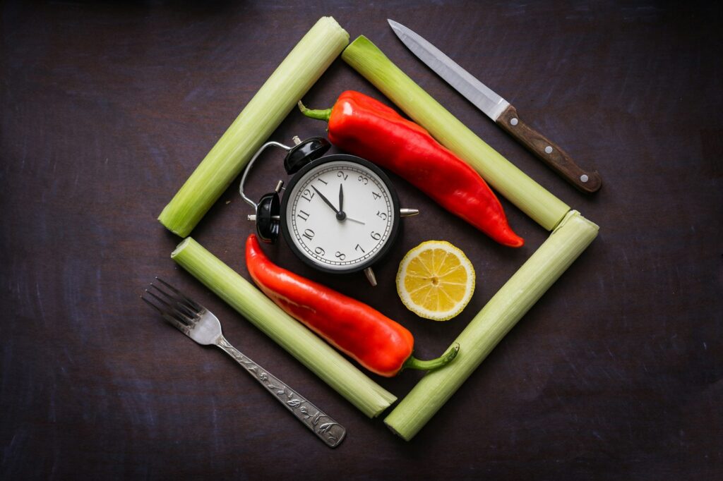 Alarm clock surrounded by vegetables on a dark table, representing meal timing.