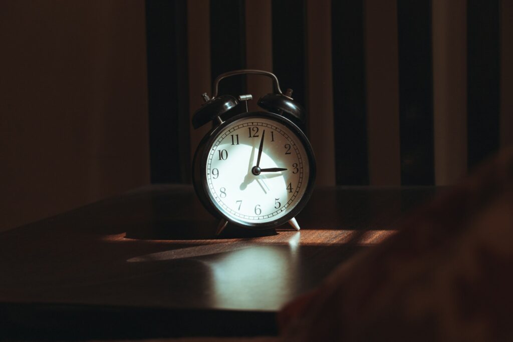 An analogue alarm clock on a bedside table in low morning light