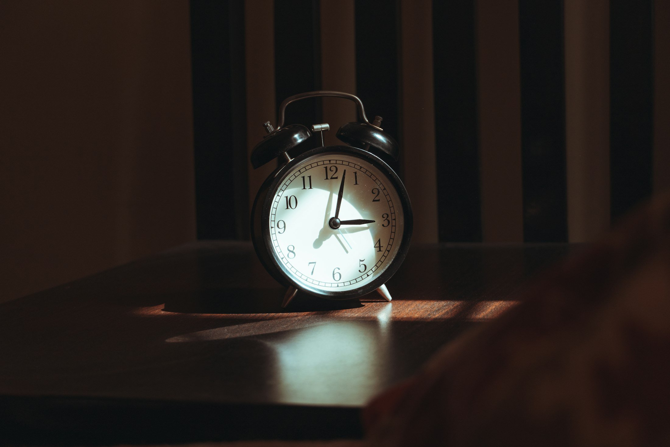 An analogue alarm clock on a bedside table in low morning light