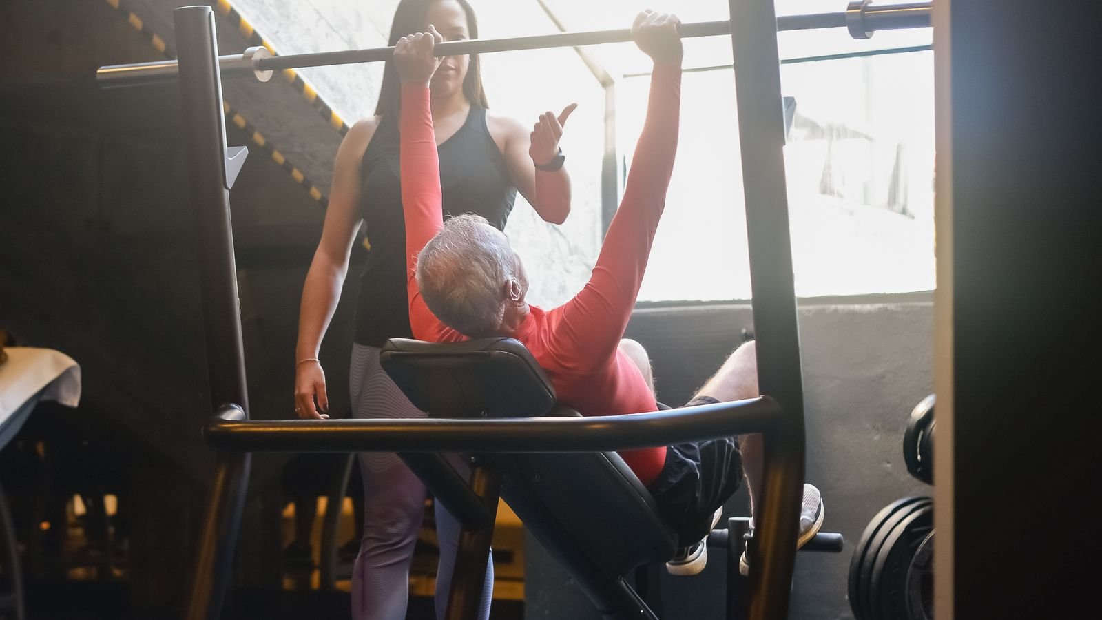 Older man lifting a barbell with a trainer in a gym