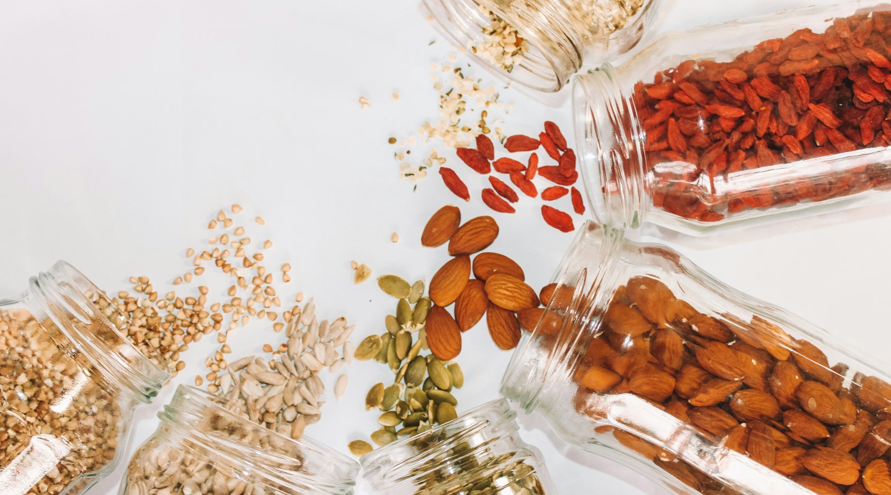 Jars of magnesium-rich foods including nuts, seeds and grains spilled on a white surface