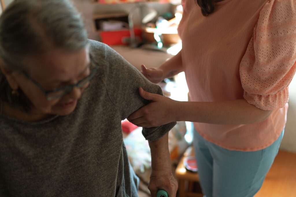 A person supports an older woman at home.