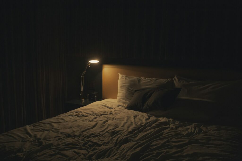 A bedside lamp illuminates pillows and rumpled bedding in a dark bedroom.